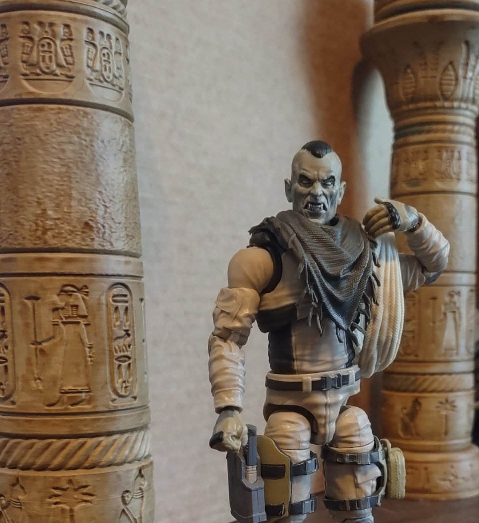 A vampire action figure stands in an Egyptian temple or possibly a tomb. He's wearing a tan uniform, thigh holster, a dull green shemagh, and has a large coil of rope on one shoulder.
