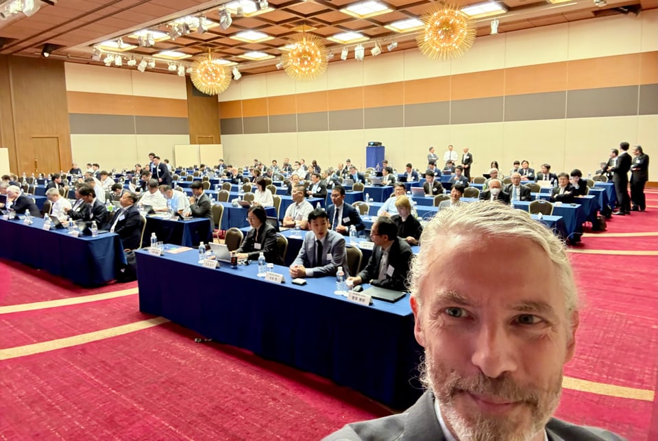 selfie of white man in suit & tie in front of a conference hall filled with business people at rows of tables