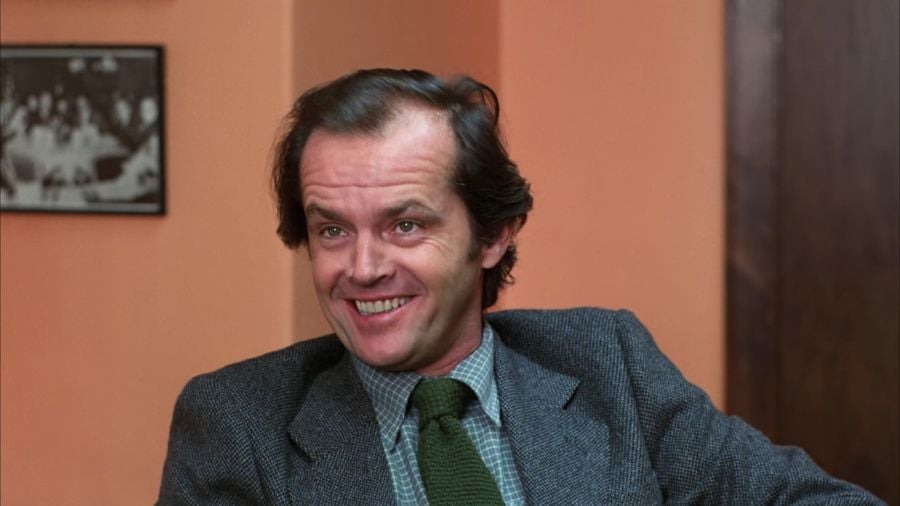 Jack Nicholson as Jack Torrance, smiling creepily, wearing a grey suit and green tie.
