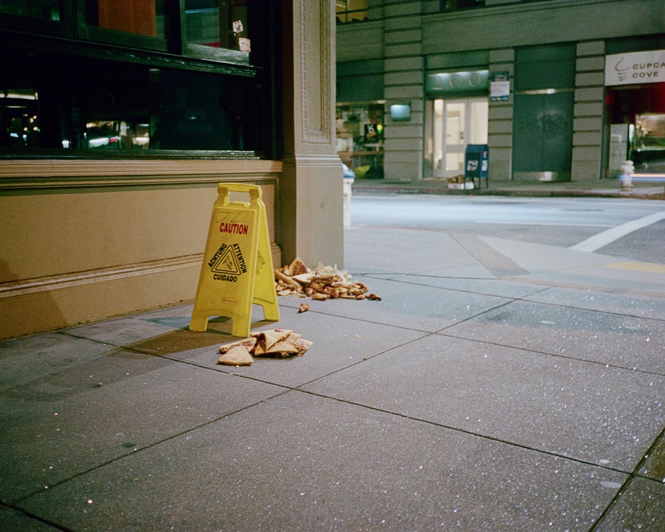 shot of a sidewalk at night, next to a building. the corner is littered with small piles of pizza. someone has placed a caution sign in their midst