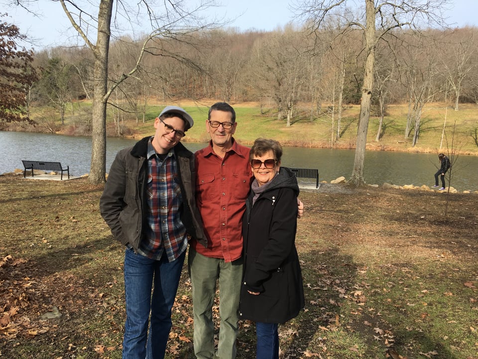 An adult child, dad, and mom stand together outside in fall in front of a lake smiling at the camera.