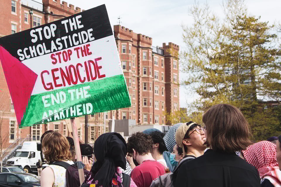 Pro-Palestine protestor holding up a sign that says "STOP THE SCHOLASTICIDE STOP THE GENOCIDE END THE APARTHEID