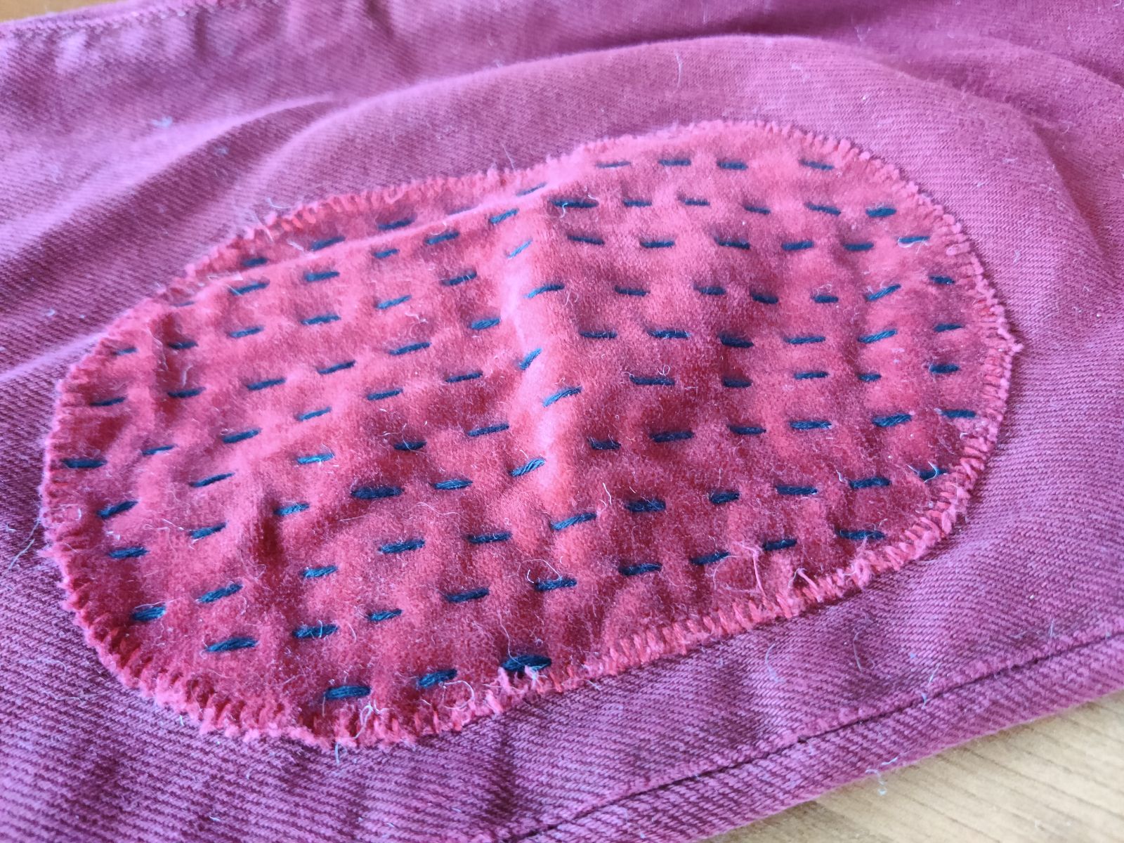 Close-up photo of 1 leg of a pair of maroon-colored pants. On the knee of the pants is a velour patch of the same color with long black running stitches in parallel lines, vertical on the patch.