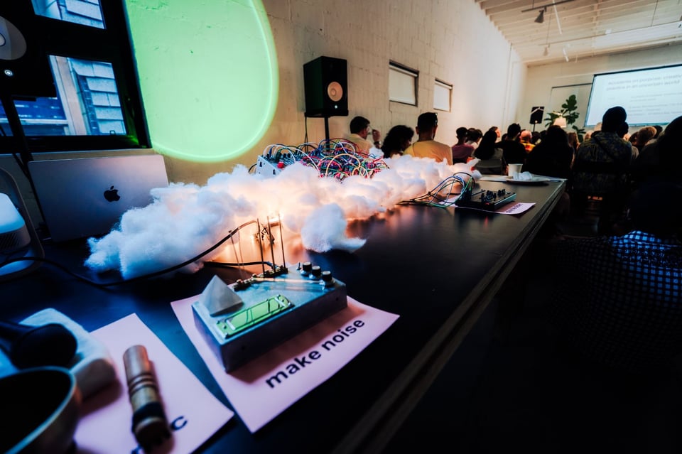 In the foreground, a synthesizer and some machinery/tools sit on a table with a sign that reads "make noise". In the background, seated people are listening to a presentation.