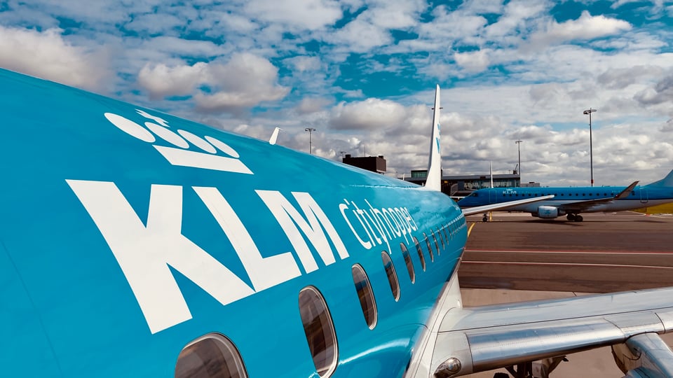 The fuselage of a KLM Cityhopper plane, with another KLM plane taxiing in the background against a KLM-blue sky dotted with small clouds
