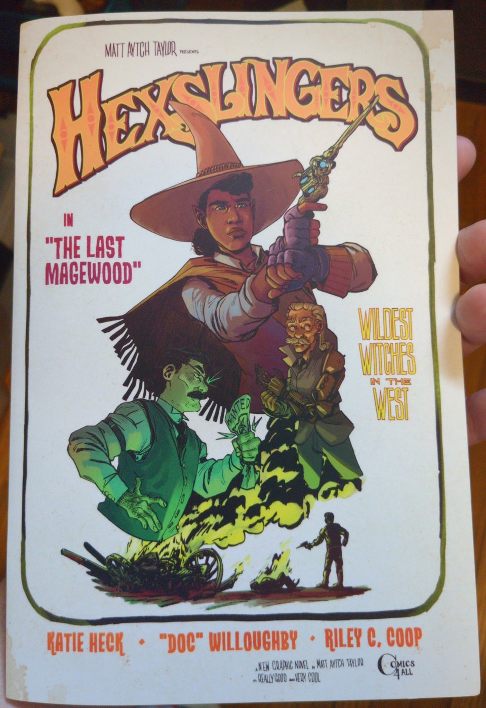 a 24 page saddle-stitch booklet containing all of my current Hexslingers preview pages, the cover is a poster-style illustration of the main characters. at the top, Katie Heck, her poncho swinging out behind her, her arms held steady wielding her six-shooter-like wand below and to the right "Doc" Willoughby holding his mech arm and looking to the side. below and to the left Riley C. Coop, lit in green clutching a wanted poster, he looks angry. and below all of them, the wreck of a horse-drawn cart, engulfed in green flame, smoke billowing out and to its right a pair of figures who have been in a fire fight.