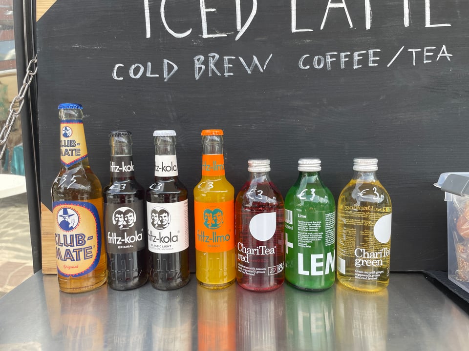A row of seven "soft drinks" in a row at a coffee truck. The most lefthand drink is "Club Mate".