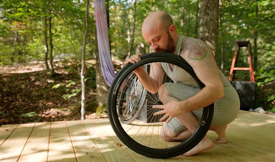 Still frame from the film: I am kneeling crouched behind a detached wheelchair wheel, with its tire tucked against my shoulder, plucking its spokes like a harp. Behind me is sun-dappled woods and a number of other objects, including more wheelchair parts.