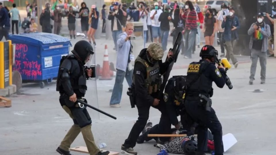 Images shows four officers assaulting a single protestor on the ground. The police are in body armour and brandishing weapons.