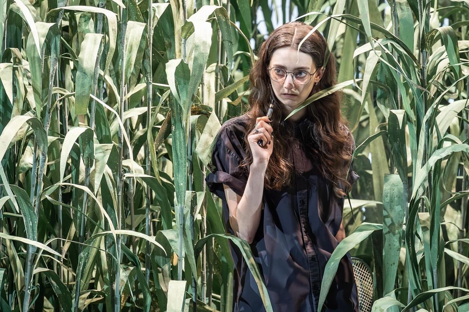 Tanya Reynolds in Thomas Ostermeier and Duncan Macmillan's The Seagull. Reynolds has long brown hair, is wearing tinted rimless glasses, a baggy black shirt and a side-bag. She is holding a vape and standing in a cornfield.
