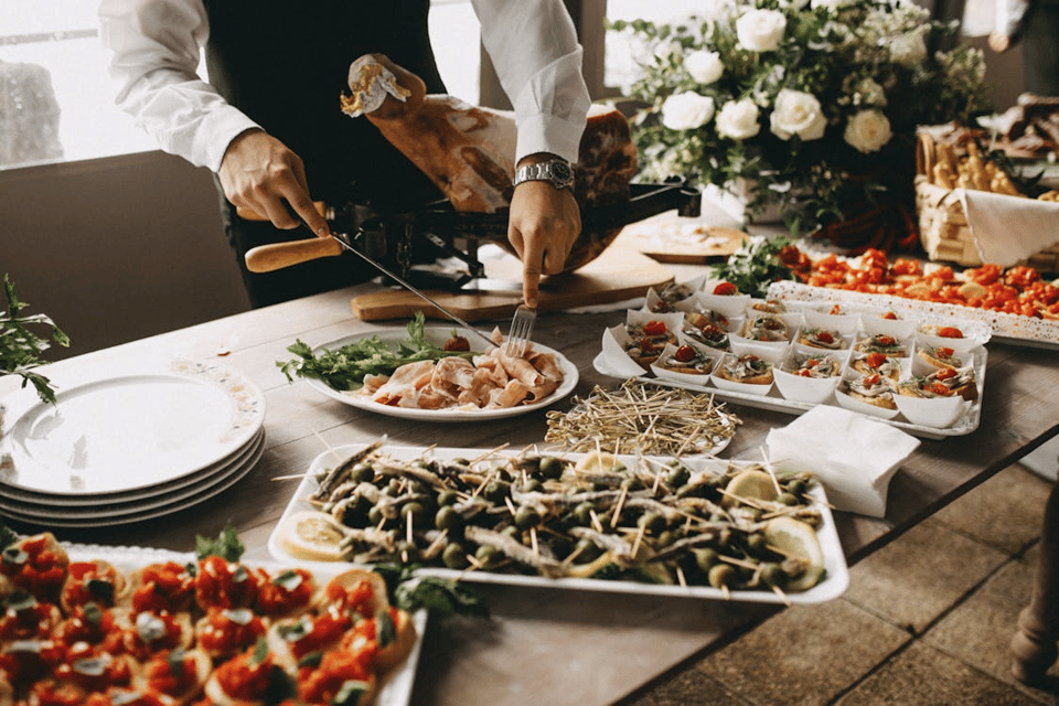 A well-dressed server is slicing cured meat at a lavish buffet table filled with an array of artfully presented appetizers. The spread includes trays of skewered olives with lemon slices, tomato-topped bruschetta, and small paper bowls filled with seafood and vegetable bites. A stack of clean white plates sits ready for guests, while a large floral arrangement and baskets of bread add elegance to the rustic wooden table. Natural light filters in, suggesting a festive and upscale gathering, perhaps a wedding or formal reception.