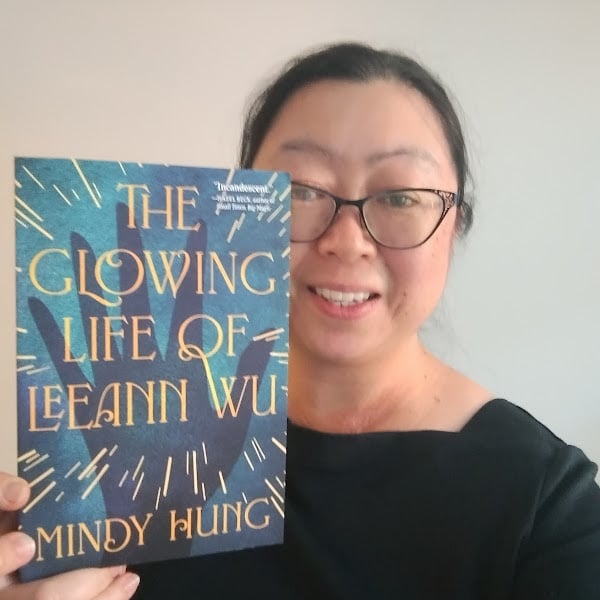 Selfie--smiling East Asian woman with slightly messy dark hair pulled back, eyeglasses--holding a paperback of the book (blue cover dark blue hand and gold sparks flying from it, gold serif title and author name) at a slight angle.
