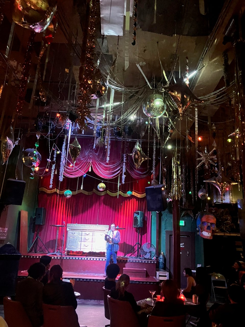 A woman reads on the stage of a dive bar decorated with silver balloons, twinkling lights, and a disco ball.