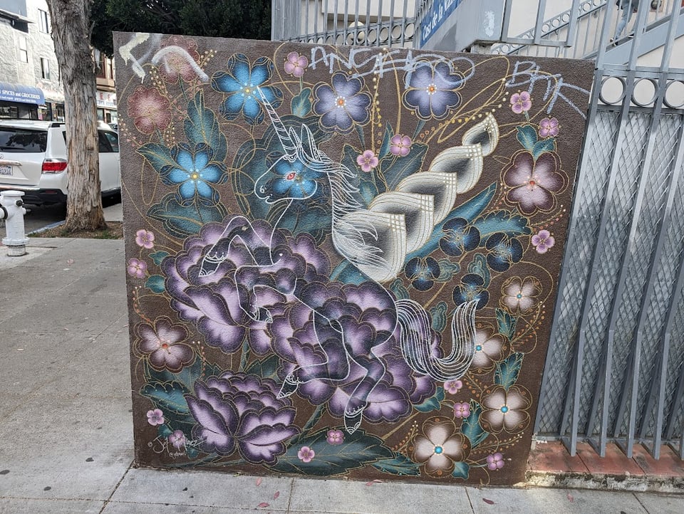 A piece of street art featuring a transparent unicorn over purple and blue flowers, plus some stalks of vegetation and silvery leaves. Everything is very sparkly.