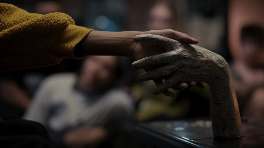 A screenshot from the film Talk To Me, showing a teenage girl's hand reaching to grasp a severed hand that is standing on a table. The hand is grey and covered in writing. The girl has brown skin and a mustard coloured jumper. Blurred figures watch in the background.