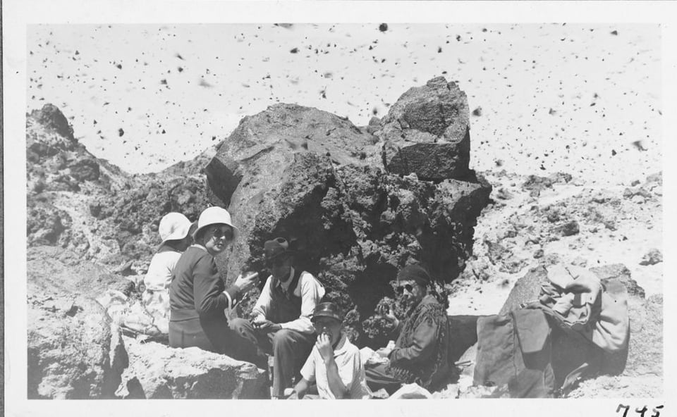 A black and white photo from the 1930s of a small group of people wearing round sunglasses in a dramatically rocky area, a few looking at the camera. The sky is open and there are thousands of black flecks in the photograph, each of which is a tortoiseshell butterfly.