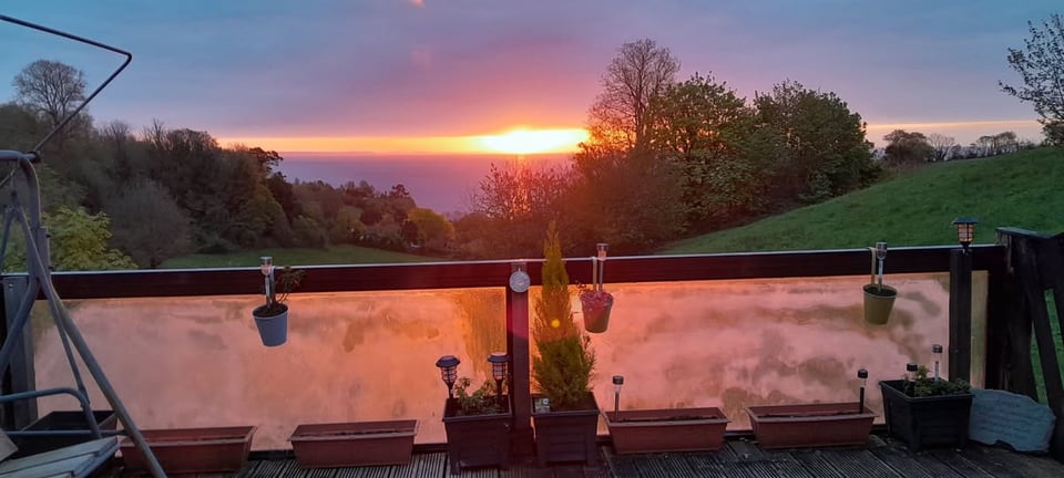 The morning sun breaking through the clouds above the sea, visible at the bottom of a steep valley. In the foreground is a balcony with frosted glass inside dark wooden frames. Planters line it. It must have been a lovely day.