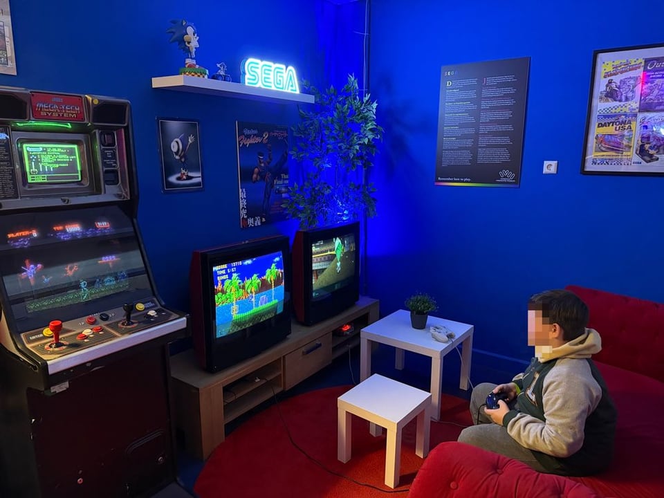A child (face blurred) plays OldSkool Sonic at the Video Game Museum in a room fashioned to be a vintage living room with a red couch and two old big box TV 📺 sets