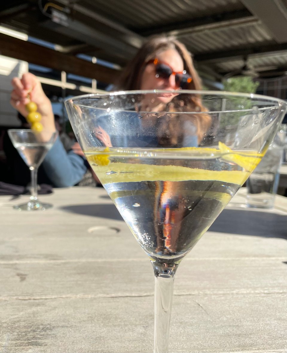 A martini in the foreground. A woman is in the background wearing shades and also drinking a martini. She has a mysterious vibe