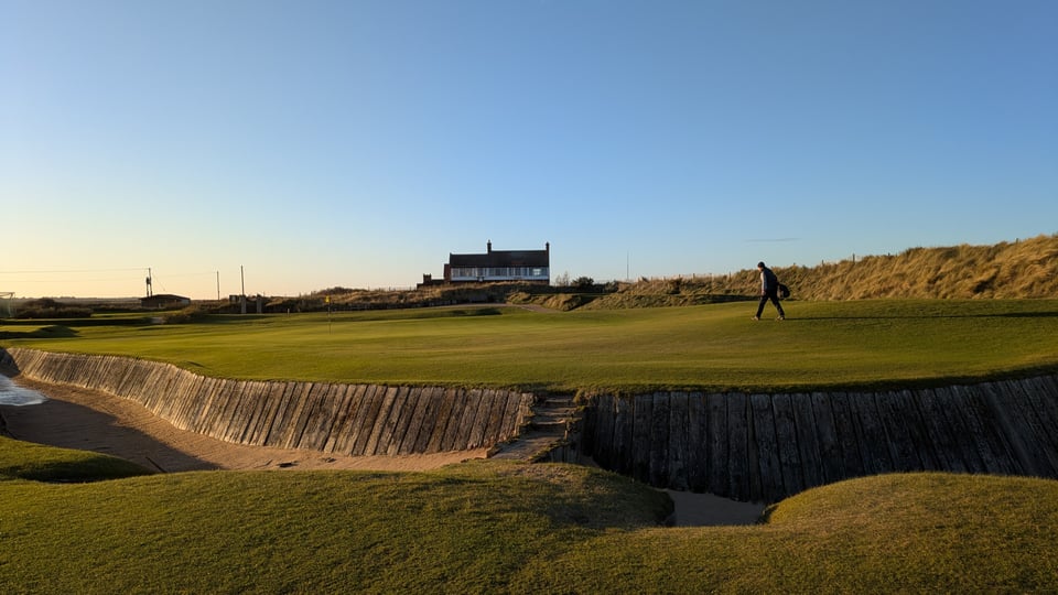 Royal West Norfolk 18th green, small bridge connecting the fairway to the green over sleeper-faced front perimeter bunker, sand dunes right, clubhouse behind, long shadows