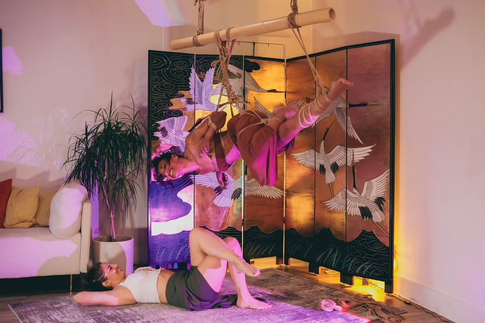 A woman (Ariana) is laying on her back on the floor in a living room, legs bent, one crossed over the other, arms behind her head as she smiles up to another woman (Euni) who's suspended in ropes from bamboo. The woman in ropes is smiling, with her arms bent and tied in rope, body slightly twisted, one leg tied straight out to the side with rope up her calf, and the other leg on top bent and tied to the straight leg.