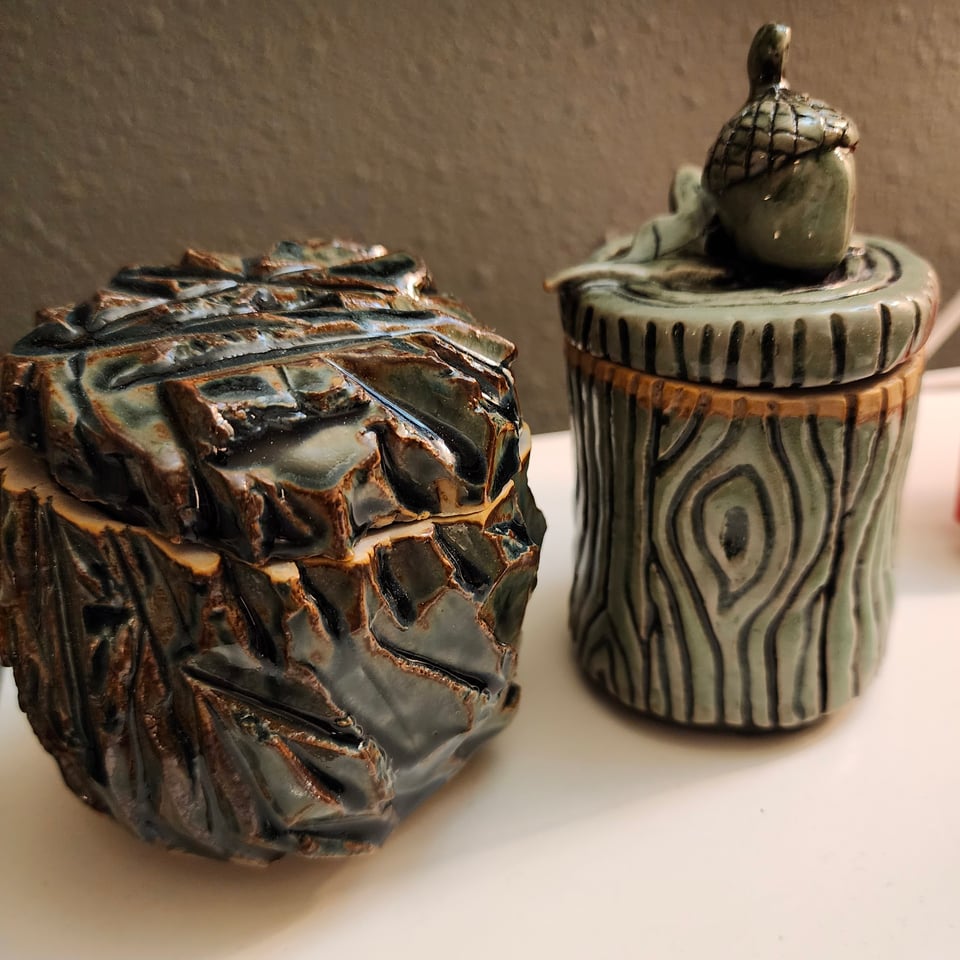 Two ceramic jars. The one on the left is rough hewn, like a craggy cliff face. The one on the right has been carved to look like wood bark. The lid has an acorn and leaf. Both of them have lids that do not quite fit.