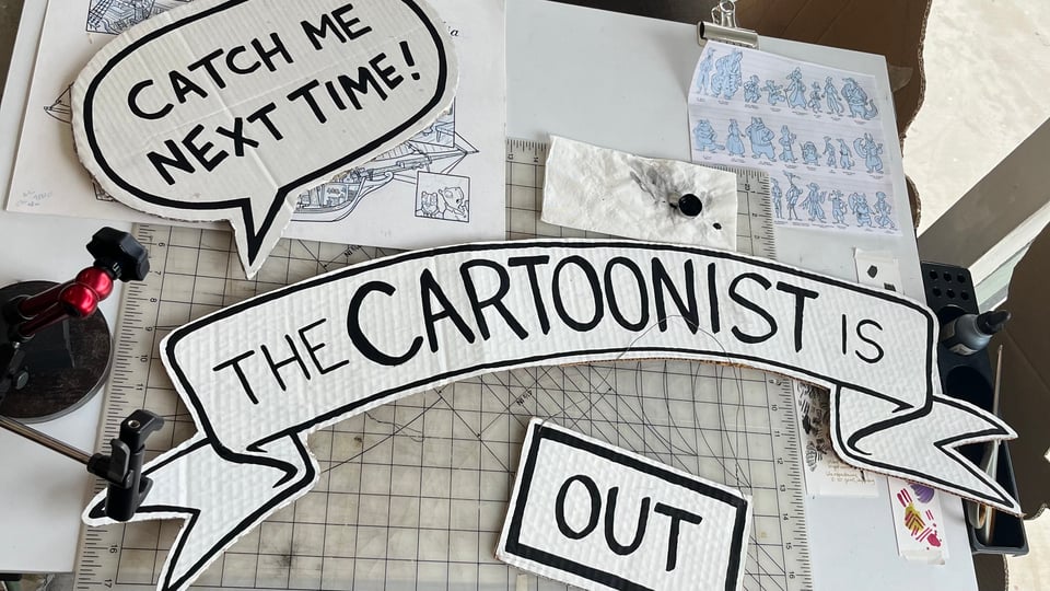 Lucy's window signage from her studio laid out on the drawing board. It reads "Catch me next time! The cartoonist is out."