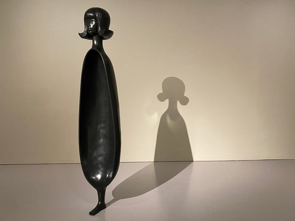 A large abstract black bronze scultpure of a woman, and the shadow it creates on the wall behind it