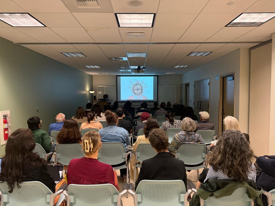 Long, narrow room set up with lecture-style seating: a full house of people facing a digital screen showing the head and shoulders of a little girl with a hairy face and wearing a brightly patterend outfit. The speaker (it = me) is on the left at a podium, mostly obscured in low lighting.