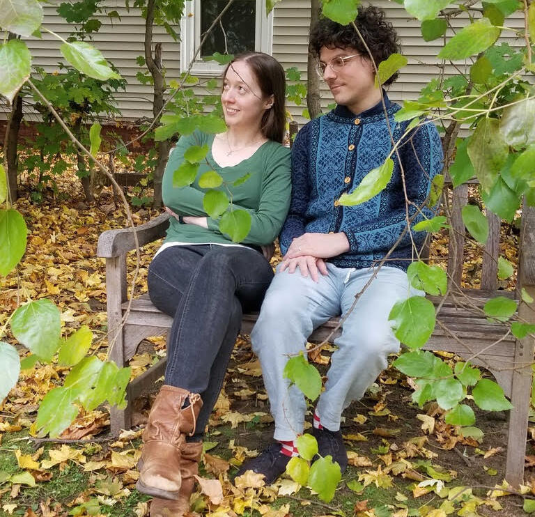 A woman in a green shirt, black jeans and light brown boots sits on a bench with a man with curly hear and glasses wearing a blue patterned sweater and light jeans.