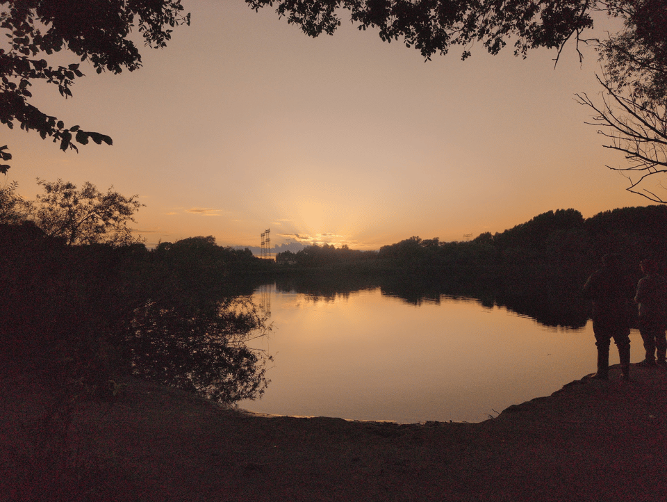 The sun is setting behind the treeline on the opposite side of a lake. The photo is mostly silhouettes of trees and a peach-colored sky. There are two people on the right side of the photo, but they are also silhouettes. The lake is still and reflecting the peach color of the sky.