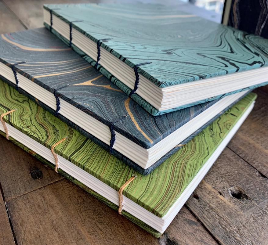 Three handcrafted notebooks with marbled covers in turquoise, dark blue, and green, stacked on a wooden surface.