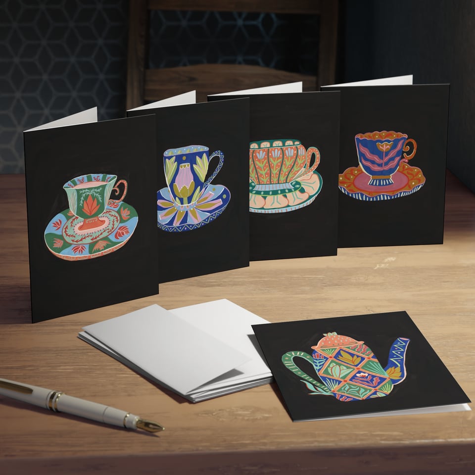 5 black greeting cards with colorful teacups and a teapot on a desk with a stack of whit envelopes and a pen