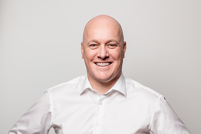 A bald white man, in a white shirt, against a white wall smiles a joyless smile. There is no life in his eyes, just a void where a person should be. This man will soon rule Aotearoa / New Zealand.