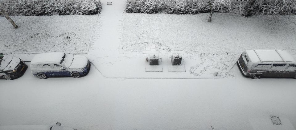 Image Description: a top-down view of a short stretch of snowy street, with a few snow-covered cars. There is grass, hedges, and a couple trees on one side of the street. The snow is unbroken, except for a faint swirly path in the grass. end ID.
