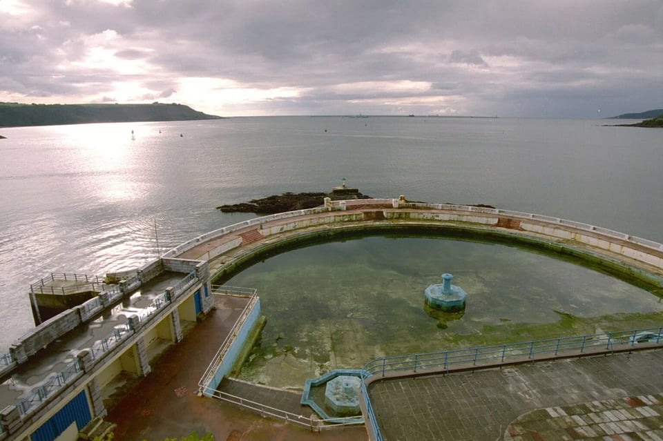 Looking over the Lido towards Plymouth Sound. The pool is half full of water, but the entirety - including the sides, walkways and fountains - is covered with green algae.