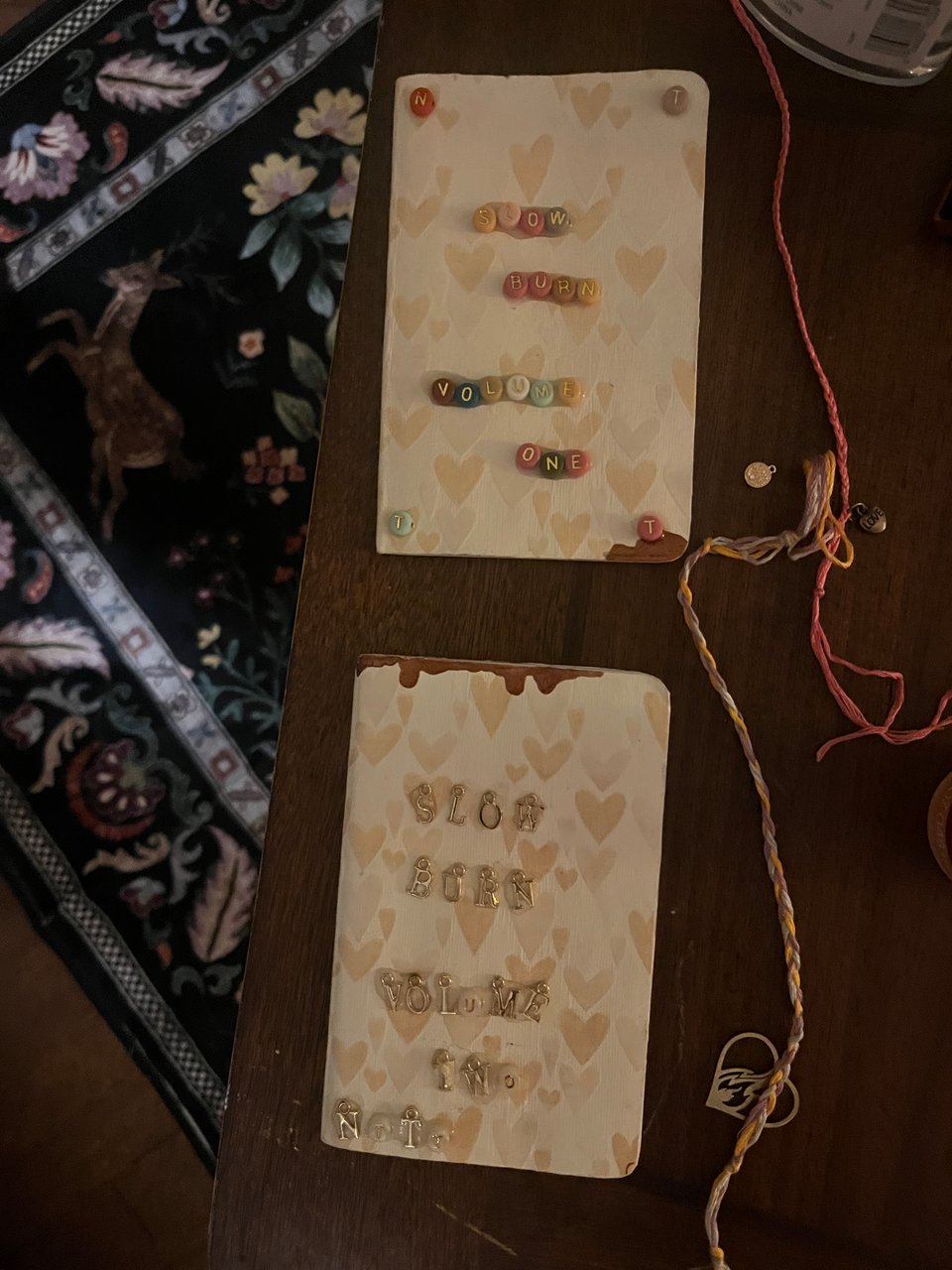Two DIY lyric books. Both are made with cream paper patterned with gold and beige hearts and have drips of dark gold wax. One says "Slow burn volume one" in multicolored round beads and the other says "Slow burn volume two" in gold charms. Multicolored friendship bracelets sit on the side, one with a gold heart charm with flames inside.