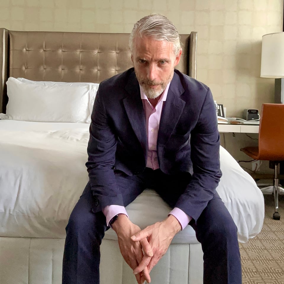 Photo of a white man sitting on the corner of a bed in a hotel room, looking at the camera, leaning forward w/forearms resting on his thighs. He's wearing a navy suit and pink shirt, with no tie.