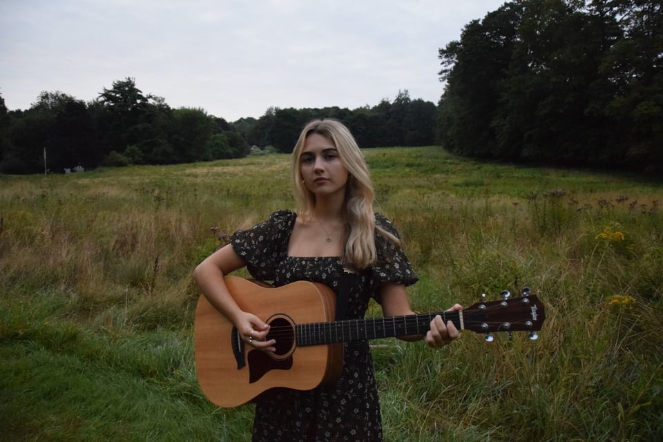 A young woman wearing a dark-colored dress, with blond hair past her shoulders, stands in a field playing an acoustic guitar.