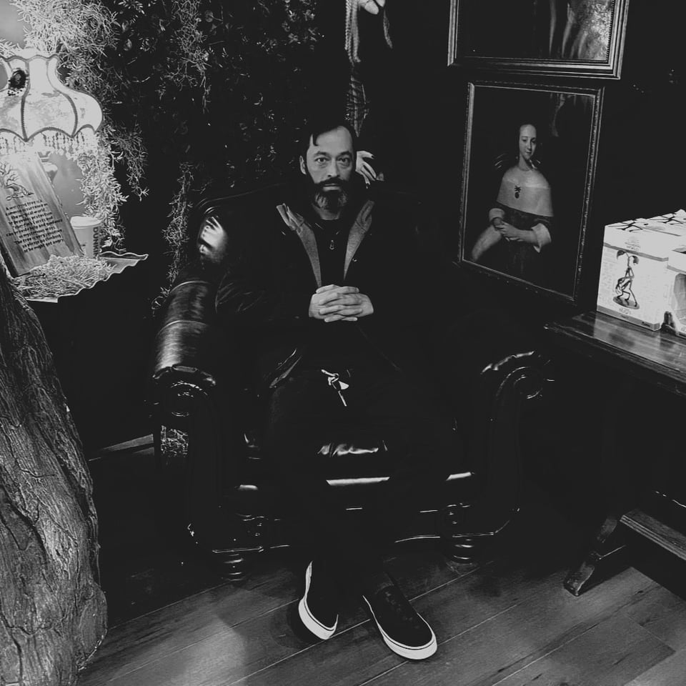 A dark black-and-white photo of man with short hair and a beard. He sits in a leather chair, hands folded in front of him, and one ankle crossed in front of the other.
