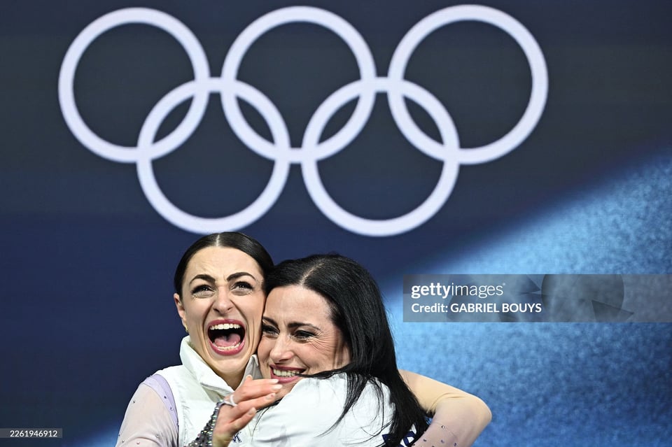 Two women embracing in front of Olympic rings, one woman screams with joy, while the other smiles.