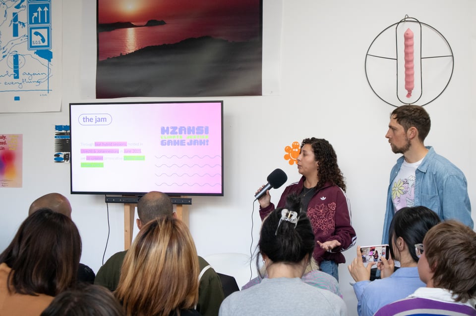 Two presenters speaking to a crowd. Screen next to them says "Mzansi Climate Justice Game Jam"