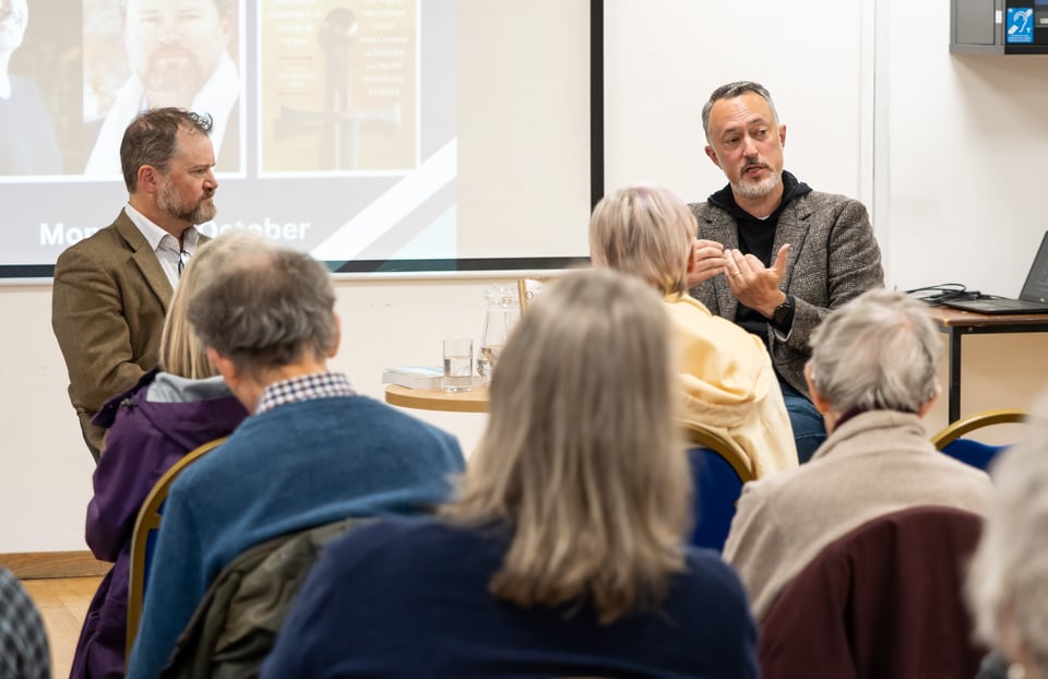 Two men sit at the front of a room; in the foreground, a bunch of people's heads seen from the back.