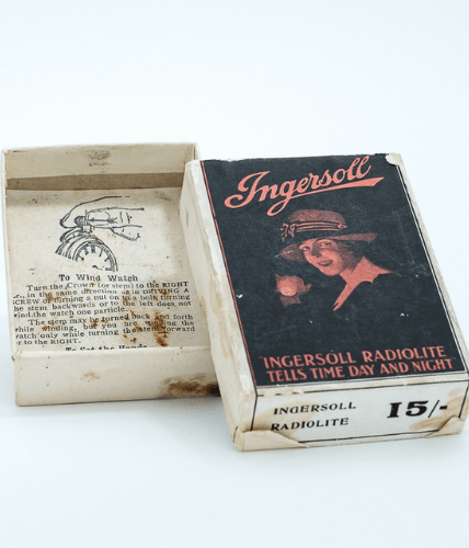 Colour photo of the top and bottom of an empty cardboard box which once contained Ingersoll Radiolite pocket watch. Top of the box has a black and orange illustration of a woman wearing the watch and smiling .Text reads 'Ingersoll Radiolite Tells Time Day and Night'