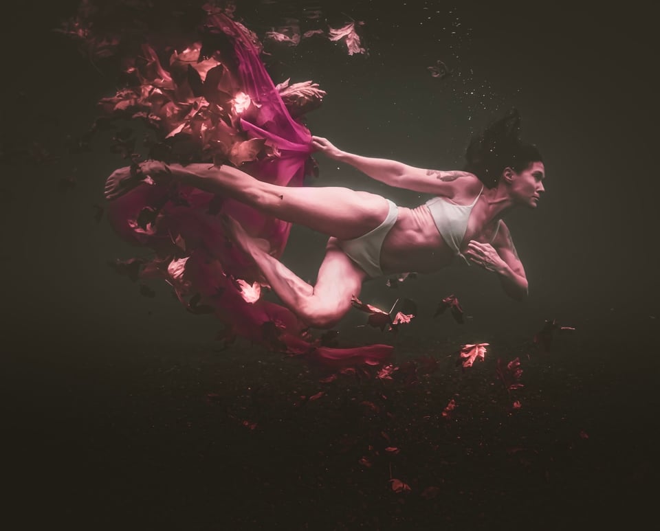 A woman in a two-piece white swimming outfit floats through dark water in the midst of swirling leaves, trailing a red fabric