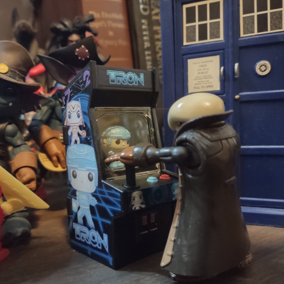 A tiny Onell Pirate Captain figure stands in front of a miniature TRON arcade game, resting one hand on the joystick. Other action figures loom behind then, along with a (relatively) huge blue police box.