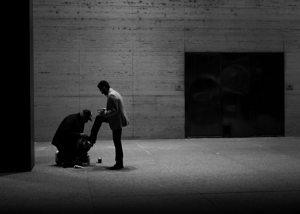 A black and white photo of a man kneeling to polish another man's shoe in a corporate plaza at night.