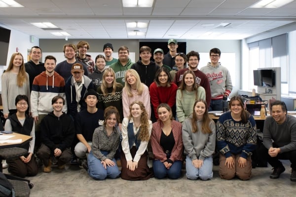 A group of students is posing for a photo inside a classroom, with some standing and others kneeling. They are dressed casually, smiling, and appear to be part of an organized event or class activity.