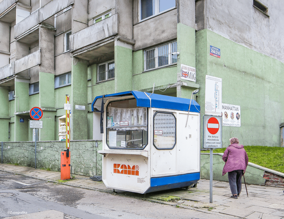 A blue and white cubic kiosk sitting on a sidewalk in front of a green brutalsit building. There is a woman in a purple winter coat and can approaching, her back to us. It is not clear what the kiosk sells