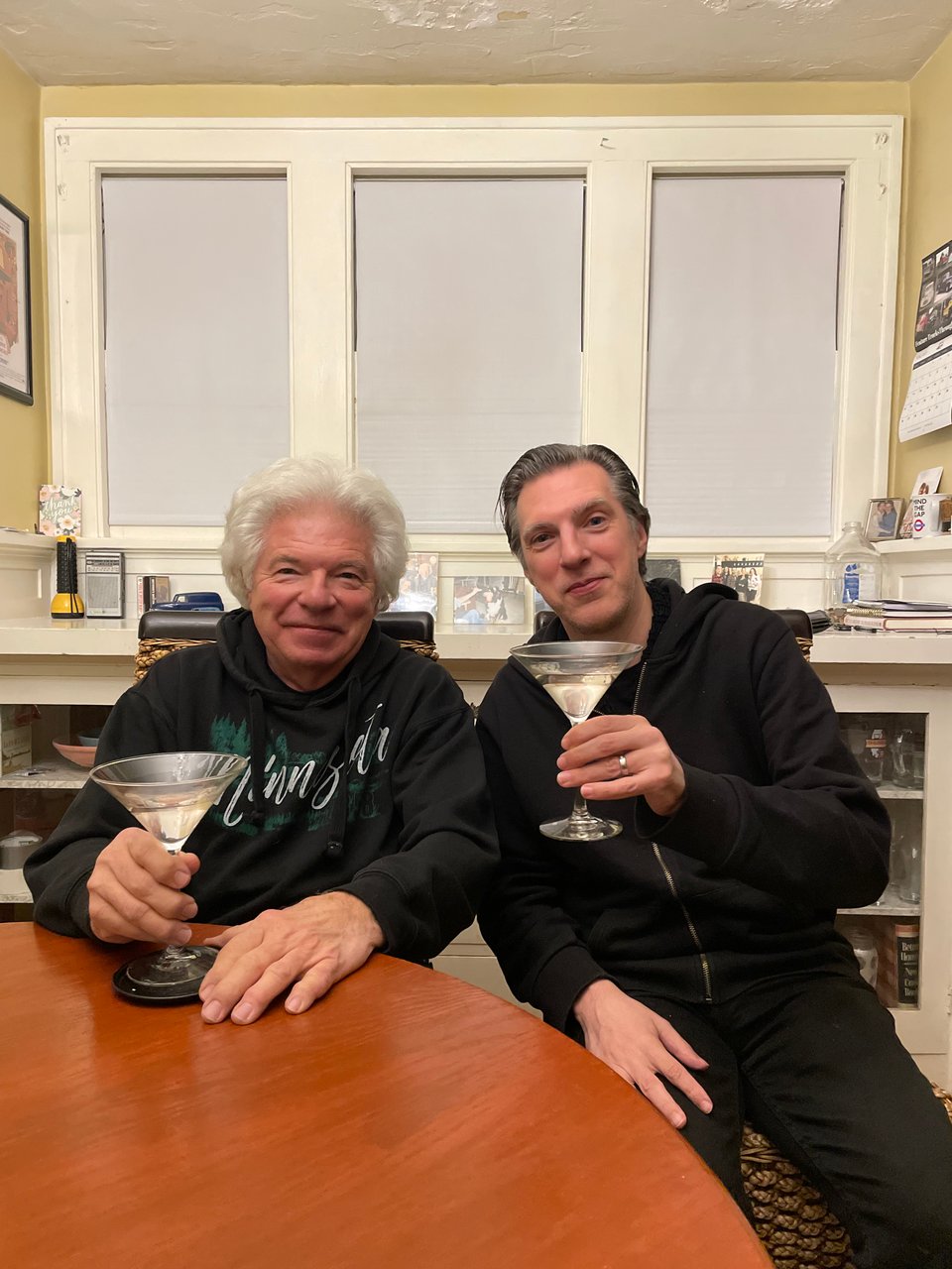 A father and a son sit in a dining room, smiling, and raising martini glasses